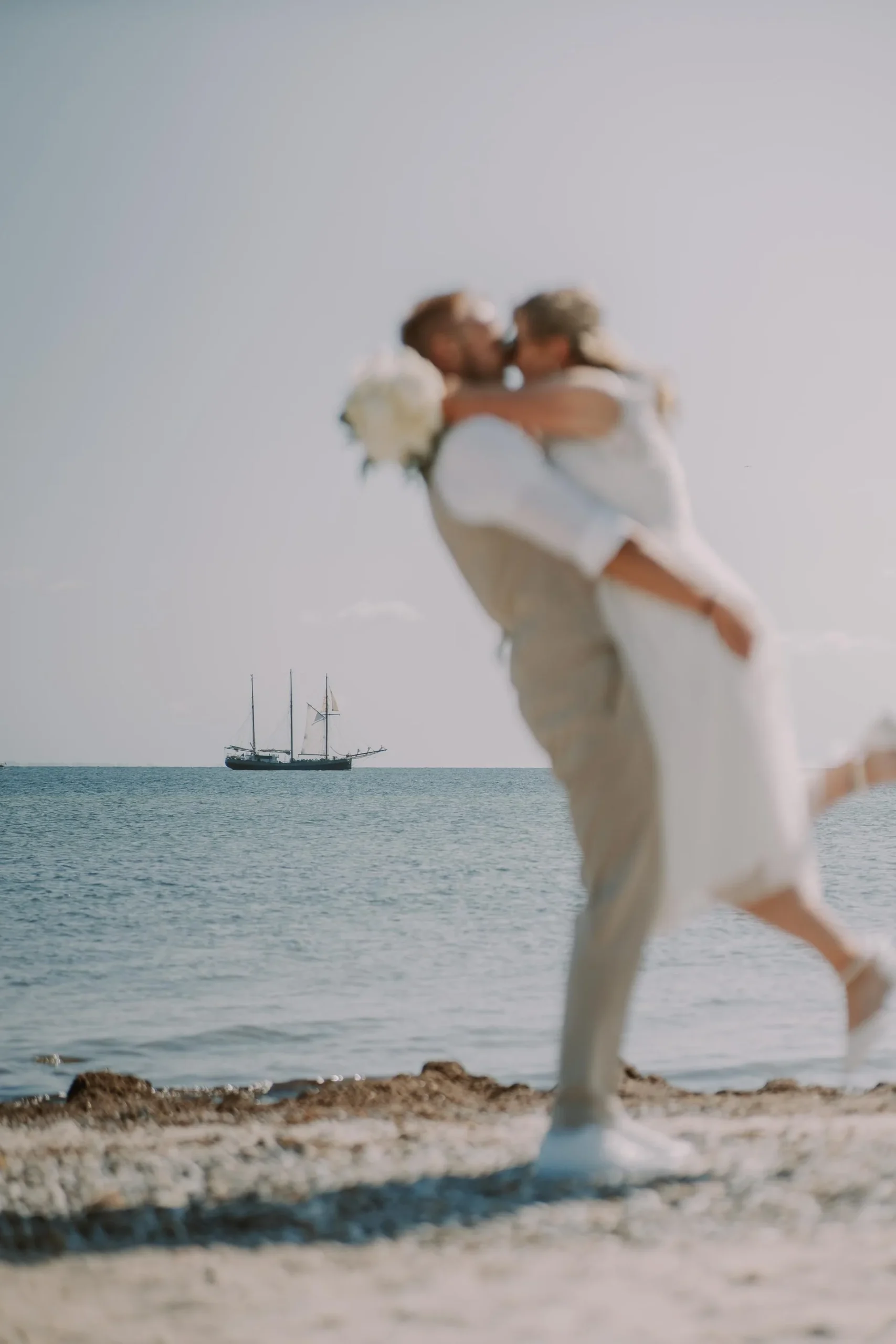 Bräutigam hebt die Braut am Ostseestrand hoch, im Hintergrund ein Segelschiff