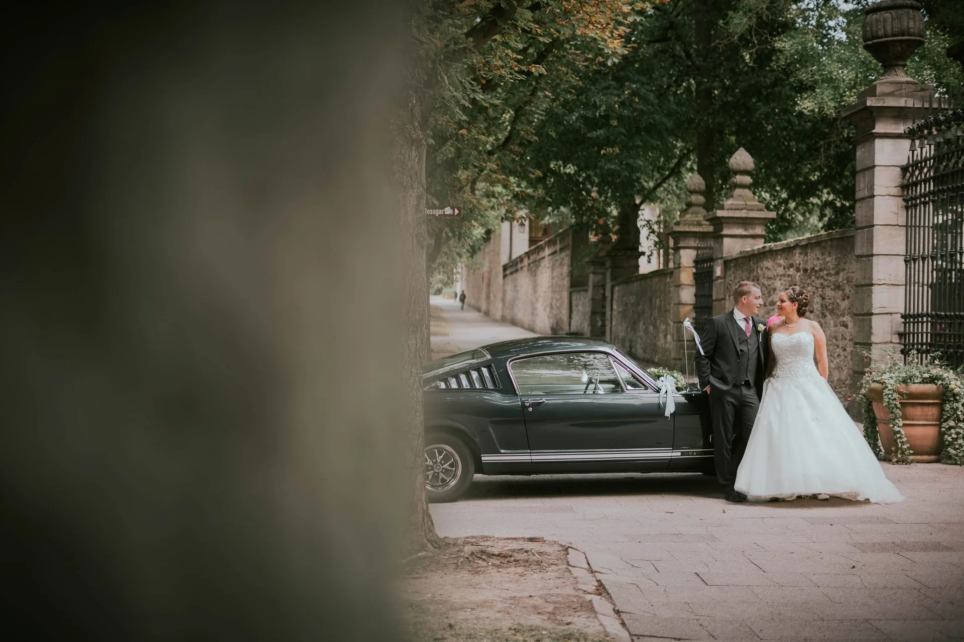 Brautpaar neben einem schwarzen Oldtimer, durch eine Säule fotografiert