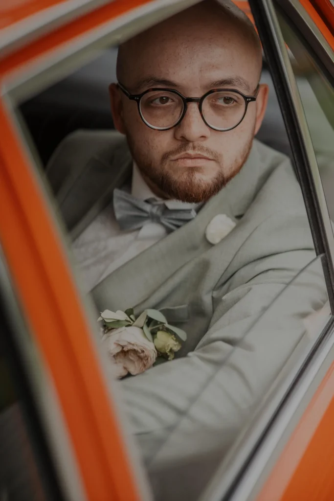 Bräutigam mit Brille sitzt in einem orangefarbenen Oldtimer in der Rhön