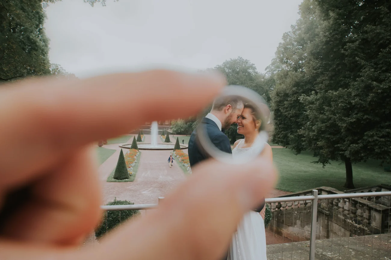 Ehering zwischen den Fingern im Schlossgarten Fulda, im Hintergrund das Brautpaar